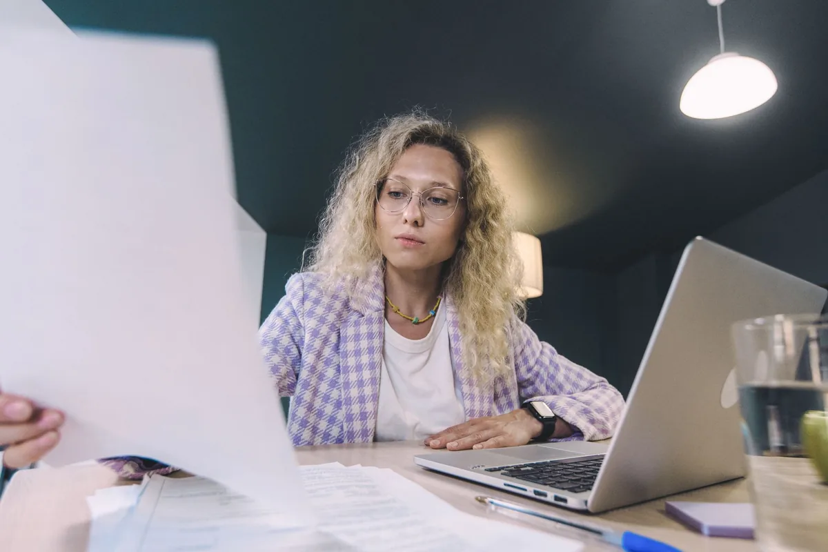 Woman in an office environment reviewing documents with focus, surrounded by technology.